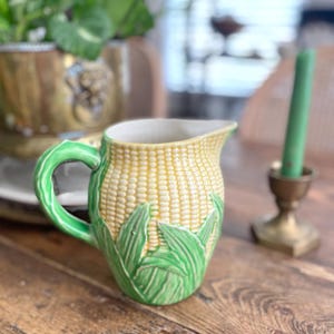 May include: A ceramic jug with a corn-on-the-cob design. The jug has a yellow body with a textured pattern, green leaves, and a green handle. A green candle and brass candlestick are in the background.
