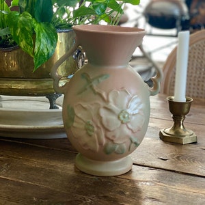 Könnte beinhalten: Eine hellrosa Keramikvase mit Blumenmuster. Die Vase hat zwei Henkel und einen erhöhten Sockel. Das Blumenmuster zeigt eine große weiße Blume mit rosa Mitte und grünen Blättern.