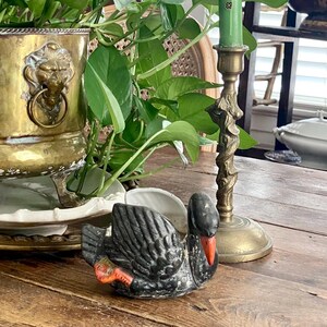 Puede incluir: Una maceta de latón con asas de cabeza de león, una vela cónica verde en un candelabro de latón y una pequeña figura de cisne negro con pico y patas naranjas, situada frente a la maceta que contiene una planta de hojas verdes.
