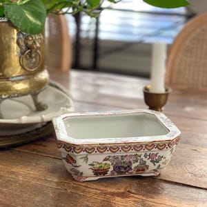 Op de afbeelding: Een rechthoekige, witte keramische plantenbak met een decoratief bloemen- en vlinderpatroon. De plantenbak heeft een rode en gouden rand en staat op een houten tafel. Een messing kandelaar en een plant staan op de achtergrond.