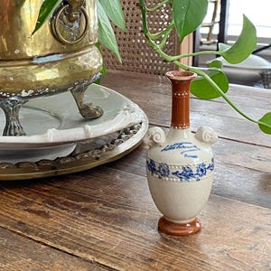 May include: A small, antique ceramic vase with a brown base and neck, and a cream-colored body. The vase features blue floral patterns and scroll-like handles. A brass planter with a lion's head handle and green leafy plant is in the background.