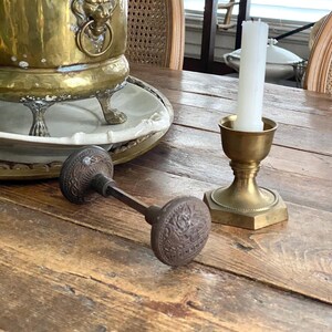 May include: A brass planter with a lion's head detail, a white candle in a brass holder, and a vintage door knob rest on a wooden table. The planter holds a green leafy plant. The table has a rustic, aged appearance.
