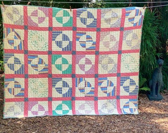 Puede incluir: Una colcha rectangular con una cuadrícula de bloques cuadrados, cada uno con un patrón circular. La tela muestra una variedad de colores que incluyen crema, rosa coral, verde azulado y azul, con pequeños estampados florales y de rayas.