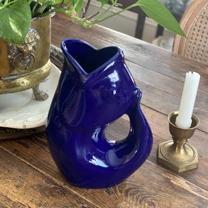 Puede incluir: Un jarrón de cerámica azul cobalto con una forma orgánica única. El jarrón tiene un acabado brillante y una gran abertura. Un candelabro de latón con una vela blanca está al lado del jarrón. El jarrón está sobre una superficie de madera.