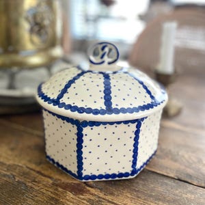 Puede incluir: Un tarro de cerámica blanco con tapa, decorado con lunares azules y bordes festoneados. El tarro tiene forma hexagonal y un pequeño asa en la parte superior. El fondo incluye una superficie de madera y una planta borrosa.