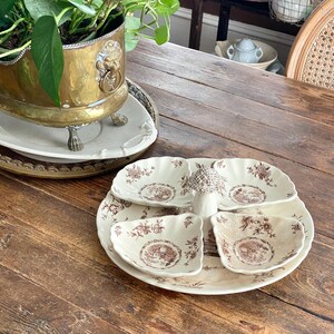 Könnte beinhalten: Eine cremefarbene Keramik-Servierplatte mit vier muschelförmigen Fächern, die um einen zentralen Griff angeordnet sind. Die Platte ist mit einem braunen Blumenmuster verziert und weist durchgehend gewellte Ränder auf.