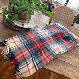 Puede incluir: Una manta plegada a cuadros en crema, rojo, verde y azul marino con bordes con flecos. La manta está sobre una mesa de madera con una planta en maceta y otros elementos decorativos en el fondo.