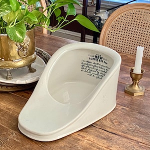 May include: A white ceramic slipper bed pan with a curved design and a wide opening. The pan has text printed on the side. A gold candle holder and a potted plant are in the background.