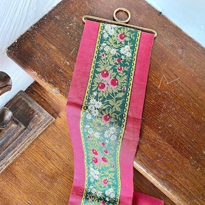 May include: A decorative wall hanging featuring a central green panel with floral embroidery of red roses and white daisies. The green panel is bordered by gold trim and framed by a vibrant pink fabric. Gold metal rings are attached at the top and bottom.