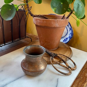 Puede incluir: Una maceta de terracota con una planta de árbol de dinero verde, una jarra de cobre y un par de tijeras tejidas sobre una superficie de mármol blanco.