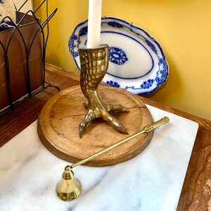 May include: A brass candle holder shaped like a bird's foot, holding a white candle. A matching brass candle snuffer rests on a wooden surface. Decorative blue and white plates hang in the background, adding a vintage touch.