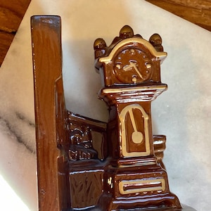Könnte beinhalten: Ein brauner Keramik-Buchstütze in Form einer Standuhr. Das Zifferblatt ist mit goldenen Akzenten versehen, und die Buchstütze hat eine glänzende Oberfläche. Die Buchstütze ist dafür konzipiert, Bücher auf einem Regal aufrecht zu halten.