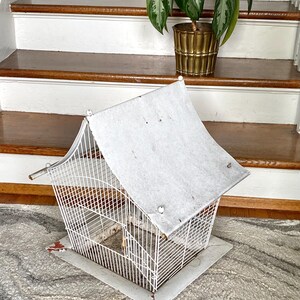 Op de afbeelding: Een vintage witte metalen vogelkooi met een puntdak. De kooi heeft een rechthoekige basis en een draadframe. Het dak is gemaakt van een lichtgekleurd materiaal. De kooi staat op een patroonkleed, met een plant in een gouden pot op de achtergrond.