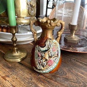 Puede incluir: Un pequeño jarrón vintage con base marrón y detalles dorados. El jarrón presenta un diseño floral en relieve detallado en tonos rosa, amarillo y verde. Tiene dos asas pequeñas y está sobre una superficie de madera. Una vela verde y un candelabro de latón están en el fondo.