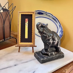 Puede incluir: Una pequeña obra de arte enmarcada con un marco dorado y un passe-partout negro. La obra de arte es una pintura abstracta vertical con colores rojo, amarillo y blanco. La obra de arte se exhibe en un pequeño caballete de madera. Una figurita de elefante negro está sobre una superficie de mármol blanco.