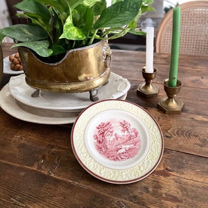 May include: A brass planter with lion head handles and claw feet holds a green pothos plant, resting on white ceramic plates. Beside it are two brass candle holders with white and green taper candles and a small plate with a red landscape print.