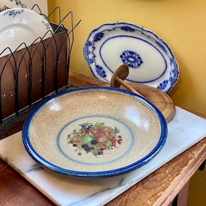 Puede incluir: Una colección de platos y cuencos de cerámica vintage. Un cuenco grande con un diseño de frutas descansa sobre una superficie de mármol. Otros platos con diseños florales azules se exhiben en la pared y en un estante de metal. La combinación de colores es principalmente azul, marrón y blanco.