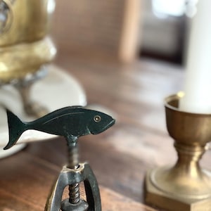 以下が含まれることがあります： 魚の形をしたハンドルが付いた、ブロンズ色のコルクスクリュー。魚には詳細なヒレと目があります。コルクスクリューのベースは、ユニークな幾何学模様のデザインです。木製の表面に置かれ、背景には他の装飾品があります。