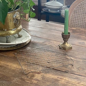 May include: A rectangular metal cooling rack sits on a weathered wooden table. A brass candle holder with a green candle is to the right. A brass planter with a lion's head design and a green plant is in the background.