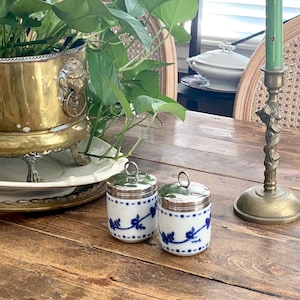 May include: Two small white ceramic jars with blue floral patterns and silver-toned metal lids featuring circular handles. The jars are cylindrical with a smooth finish, showcasing a delicate vine design in blue ink encircling the exterior surface.