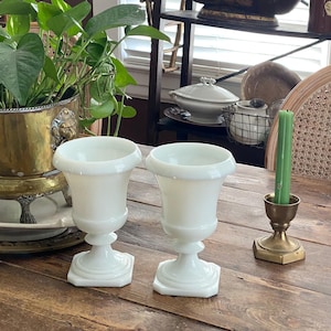 May include: Two white, vintage-style goblets with a pedestal base and flared rim. The goblets are made of opaque glass and sit on a wooden table. A green candle in a brass holder is to the right.