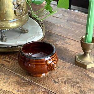 May include: A small, round, brown ceramic bowl with a glossy finish and a molded lion head detail on the side. The bowl features a dark rim and sits on a wooden surface alongside a brass candlestick holding a light green taper candle.