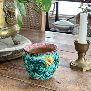 May include: A small, textured ceramic pot with a turquoise and green exterior, a pink and white rim, and a yellow flower design. The pot sits on a wooden table next to a brass candle holder and a larger brass planter.