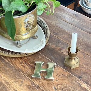 May include: A brass planter with a lion's head detail, a white candle in a brass holder, and a metallic letter "H" sit on a wooden table. A white ceramic plate and a decorative tray are also visible. The scene has a vintage aesthetic.