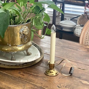May include: A brass electric candle with a cream-colored candle and a clear bulb. The candle sits in a brass holder on a wooden table. A brass planter with a lion's head design and green plants is in the background.