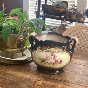 Könnte beinhalten: Ein antiker Keramik-Blumentopf mit gewelltem Rand und zwei Griffen. Der Topf hat eine cremefarbene Basis mit einem Blumendesign in Rosa- und Lilatönen. Der Rand und die Griffe sind dunkelbraun.