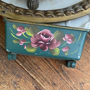 Peut inclure: Petite jardinière rectangulaire en métal vert, ornée d'un motif floral peint à la main. Les fleurs sont bordeaux foncé avec des accents roses et des feuilles vertes. La jardinière a de petites roues vertes.