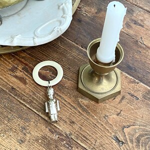 May include: A vintage arrangement showing a brass candlestick with a white candle and a silver soldier charm. The charm is suspended from a cream ring. The items are displayed on a wooden surface, creating a warm, antique aesthetic.