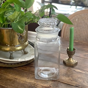 May include: A clear, square glass jar with a decorative lid. The jar sits on a wooden table, next to a brass candle holder with a green candle and a brass planter with a green plant.