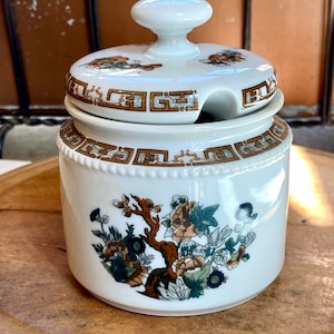 May include: White ceramic sugar bowl with a lid and a floral design. The bowl has a gold trim and a spout for pouring.