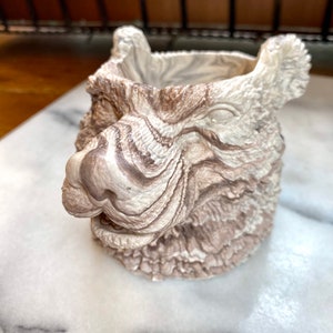 May include: A decorative planter shaped like a bear's head, featuring a textured, marbled design in shades of brown and cream. The bear's face is detailed with a nose, eyes, and ears, creating a unique and artistic piece for home decor.