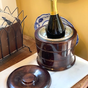 May include: A dark brown wooden wine chiller with a silver interior holds a champagne bottle with a gold foil top. The chiller sits on a white marble surface, with its matching lid beside it. Decorative blue and white plates are in the background.