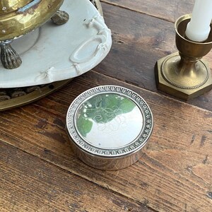 May include: A small, round silver box with a decorative beaded edge and a reflective lid. The lid features an engraved monogram. The box sits on a wooden surface, alongside a brass candlestick and a white ceramic tray.