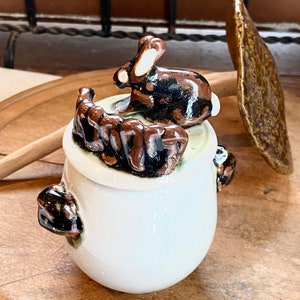 Puede incluir: Un tarro de cerámica blanco con tapa que presenta una figura de conejo marrón y negro. El tarro tiene dos asas pequeñas y está sobre una superficie de madera. Una cuchara de cerámica marrón descansa cerca. El tarro es probablemente para uso en la cocina.