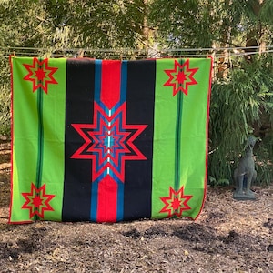 Puede incluir: Una colorida manta con un diseño de estrella geométrica. La manta presenta una franja negra central flanqueada por franjas verdes, con detalles rojos y azules. Estrellas rojas están colocadas en las franjas verdes. La manta está colgada al aire libre.