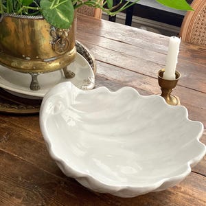 Peut inclure: Un grand bol blanc en céramique en forme de coquillage avec un bord ondulé. Le bol est posé sur une table en bois, à côté d'une jardinière en laiton avec de la verdure, d'une bougie blanche et d'un bougeoir en laiton. Le design du bol évoque un thème côtier.