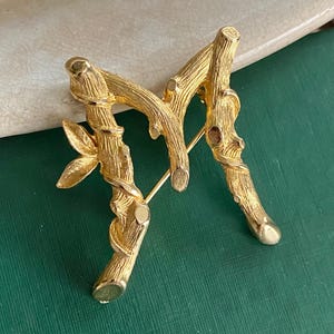 May include: A gold-toned brooch in the shape of a stylized letter "M". The brooch is designed to resemble intertwined branches with a leaf detail. The pin is set against a green background.
