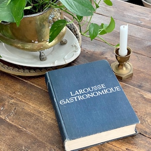May include: A blue hardcover book titled "LAROUSSE GASTRONOMIQUE" rests on a wooden surface. A brass candle holder with a white candle and a brass planter with green plants are also visible.