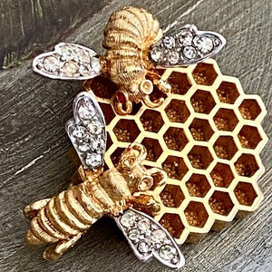 May include: A gold-toned bee brooch with rhinestone-studded wings and a honeycomb-shaped base. The bee's body has detailed ridges, and the wings are outlined in silver. The brooch is a decorative accessory.