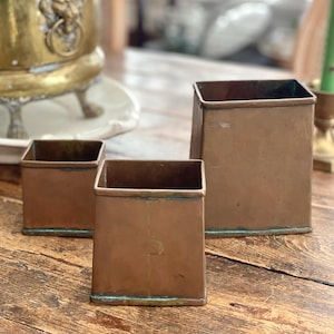May include: Three square copper containers of varying heights with a weathered patina. Each vessel features a flat base, straight sides, and an open top. The metal surfaces show signs of oxidation with visible green verdigris along the edges.