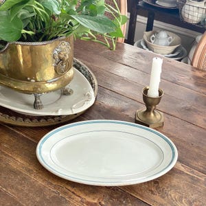 Könnte beinhalten: Eine ovale, weiße Keramikplatte mit hellblauem Rand. Ein Messing-Kerzenhalter mit einer weißen Kerze steht auf einem Holztisch. Ein Messing-Pflanzgefäß mit Löwenkopf-Design enthält eine grüne Pflanze.