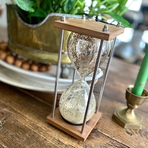 May include: An hourglass with a silver mercury glass bulb, set in a wooden frame with stainless steel supports. The hourglass is on a wooden surface, with a brass candle holder and a green candle in the background.