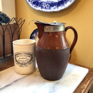 Puede incluir: Una jarra de cerámica marrón y beige con tapa y asa de metal, junto a un tarro de cerámica blanco con texto negro "James Keiller & Sons Dundee Marmalade". Un plato con estampado azul y blanco es visible en el fondo.