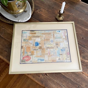 Peut inclure: Une œuvre d'art encadrée, composée d'un collage d'objets éphémères vintage, tels que des timbres et des billets. L'œuvre est encadrée dans un cadre doré et un passe-partout crème, avec une bordure bleu foncé. Le texte "THIS & CO'S" est visible.