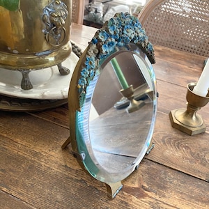 Op de afbeelding: Ovale toilettenspiegel met een messing frame en voet. De bovenkant van het frame is versierd met blauwe bloemversieringen. De spiegel staat op een houten tafel, met messing plantenbakken en kandelaars op de achtergrond.