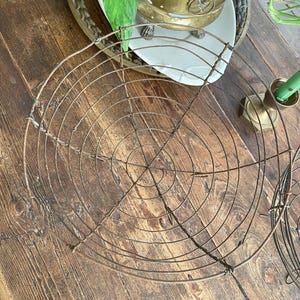 Könnte beinhalten: Ein runder, antiker Metall-Kühlrost mit konzentrischem Drahtdesign. Der Rost besteht aus dunklem, gealtertem Metall und liegt auf einer rustikalen Holzoberfläche. Das Design zeigt eine Reihe von konzentrischen Kreisen und radialen Drähten.